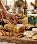 Hand cutting fresh Happy Bees Market raw honeycomb with a wooden spoon on a large party charcuterie board with brie, crackers, sliced apples, figs, grapes, and nuts.